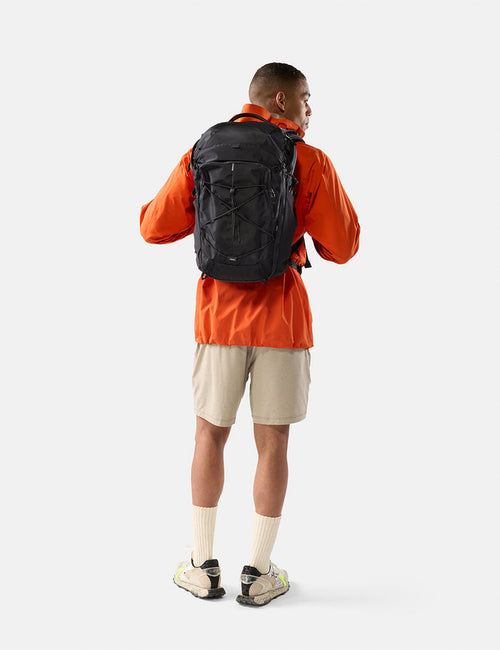 Person wearing an orange jacket and beige shorts with a black backpack on a white background