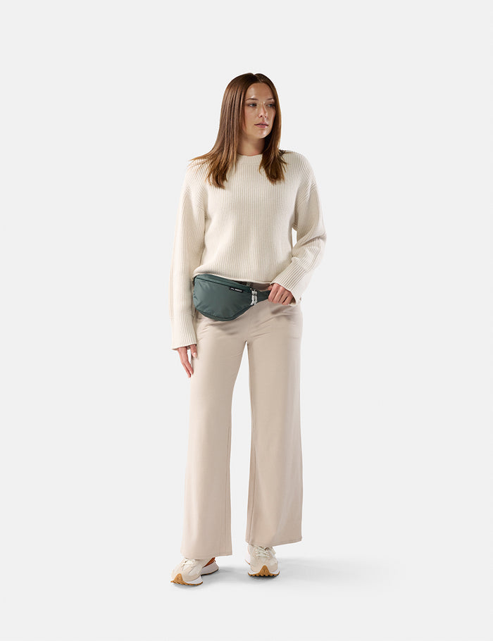 Woman wearing a cream sweater and beige pants with a green fanny pack on a white background