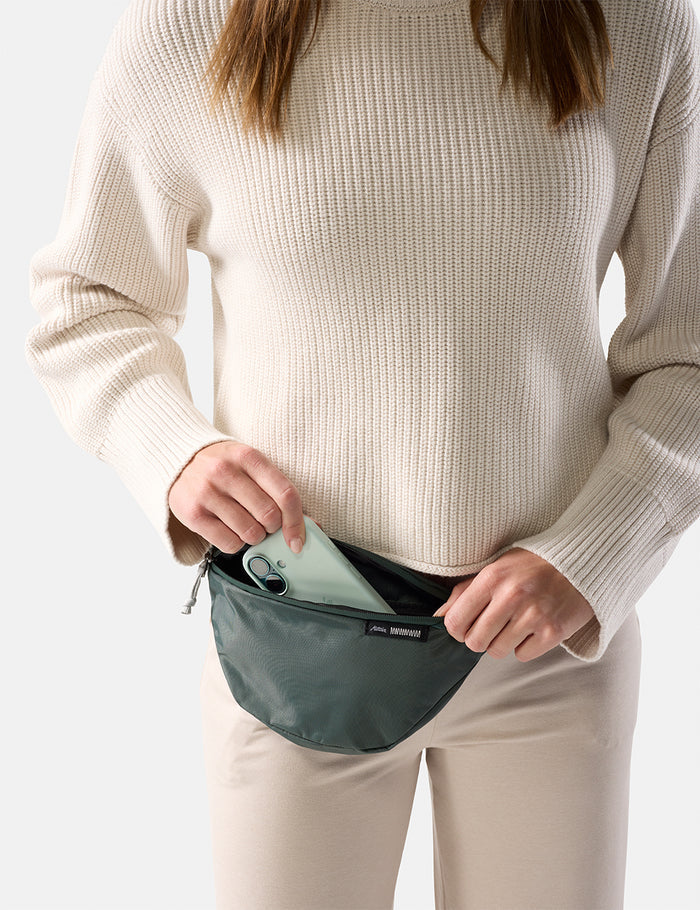 Person wearing a beige sweater and white pants, holding a teal waist bag with iphone