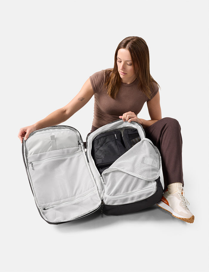Woman sitting on the floor with a large open backpack, showing its compartments.