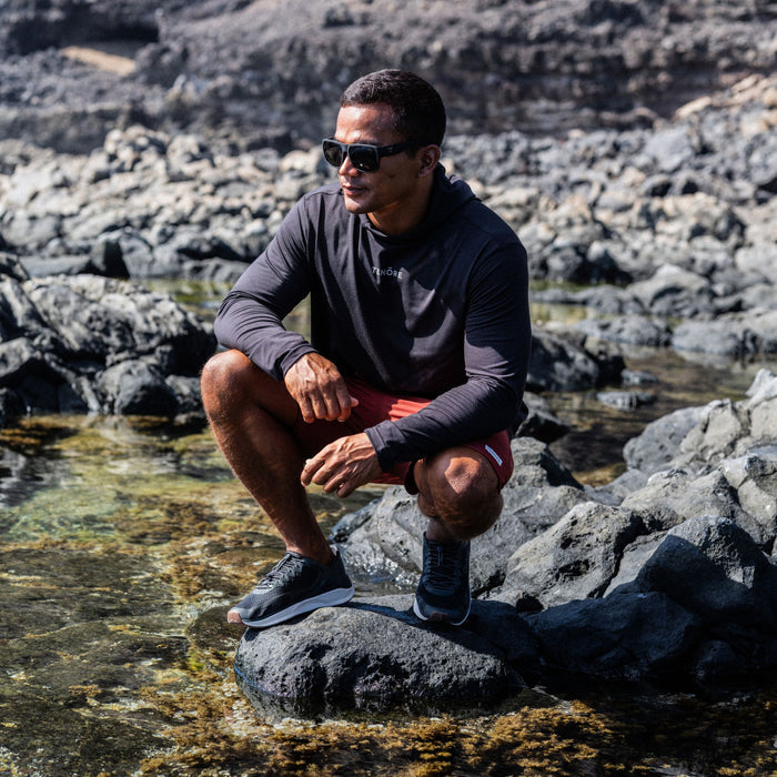 Man wearing black long-sleeve rashguard and coral shorts crouching on rocky shore with sunglasses.