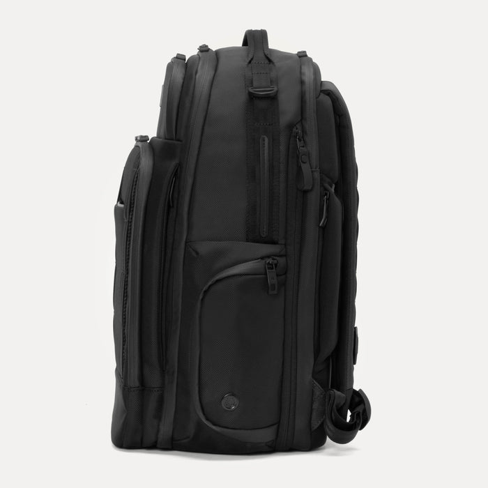 Side view of the Travelpro Altitude Large Expandable Laptop Backpack in Black, showing multiple zippered compartments, padded side panels, and the handle attachment against a light background.