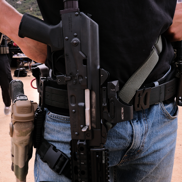 Close waist shot of a person wearing a black Kore Essentials laser cut B2 battle belt and other tactical gear with a rifle and backpack in an outdoor setting