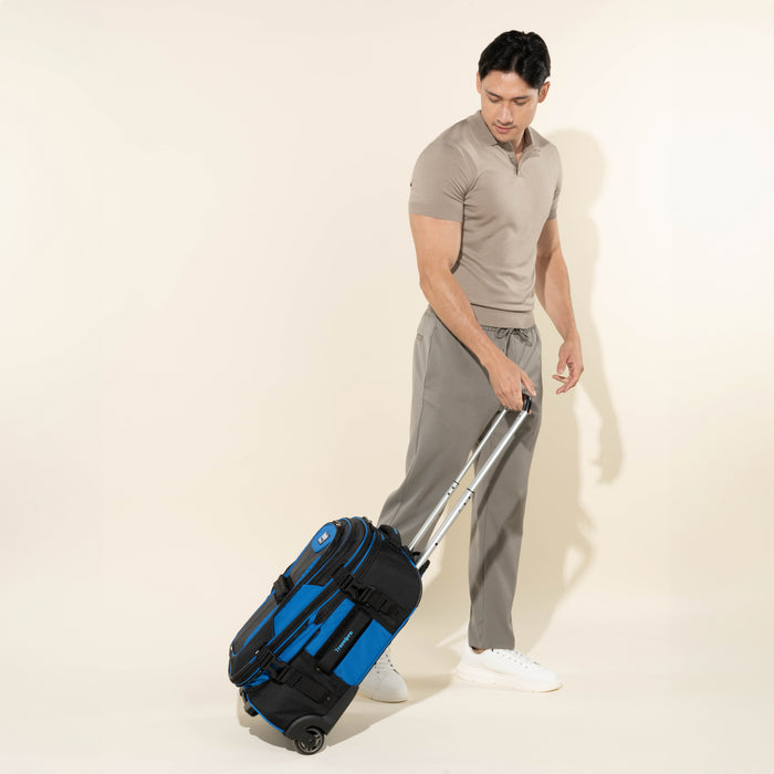 lifestyle view of a male model in a taupe polo shirt and grey pants tilting back and walking with a blue black bold by travelpro carry-on expandable rollaboard suitcase, showing the bag in motion on its two wheels with the telescoping handle extended.