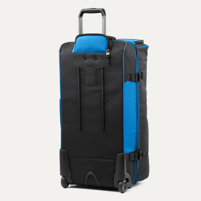 rear view of the blue black bold by travelpro large check-in drop-bottom rolling duffel bag standing vertically, showing its protective skid guards, reinforced wheel housings, two high-performance ball-bearing wheels, and extended telescoping handle.