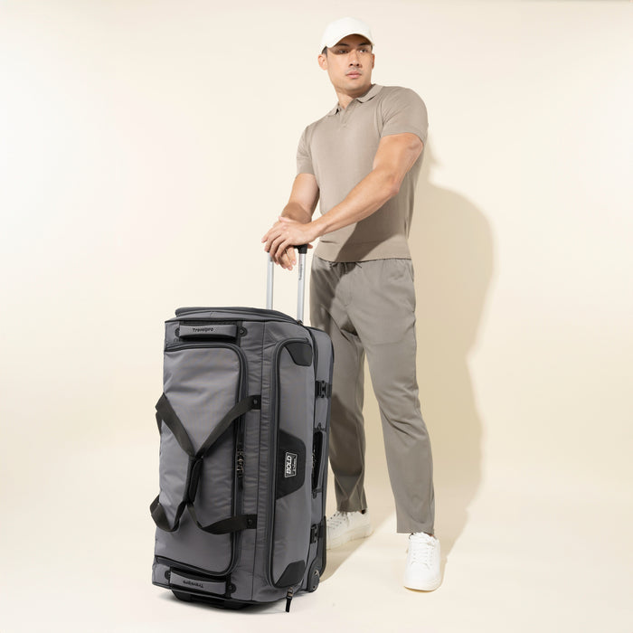 lifestyle view of a male model in a polo shirt and grey pants standing with the gray black bold by travelpro large check-in drop-bottom rolling duffel bag, showing its upright vertical scale and extended telescoping handle.