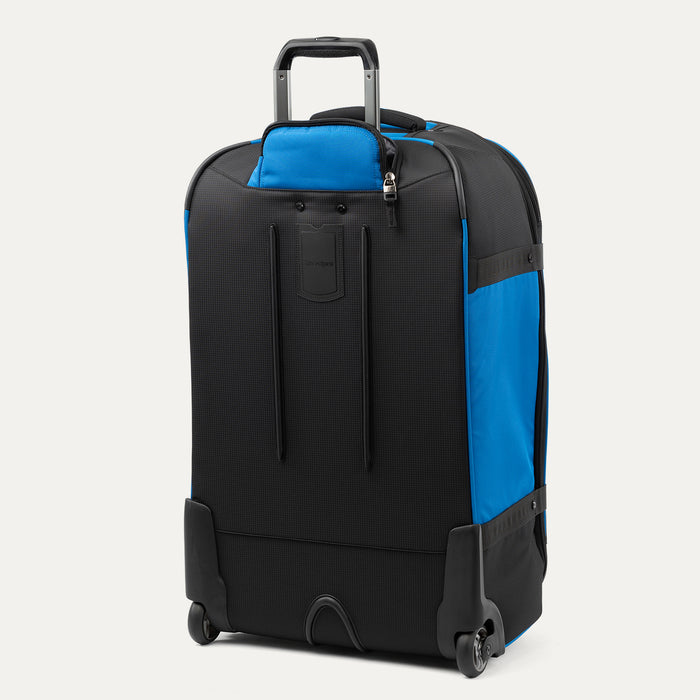 rear three-quarter view of the blue black bold by travelpro large check-in expandable rollaboard suitcase standing vertically, showing its protective skid guards, molded corner guards, reinforced wheel housings, two high-performance ball-bearing wheels, and extended telescoping handle.