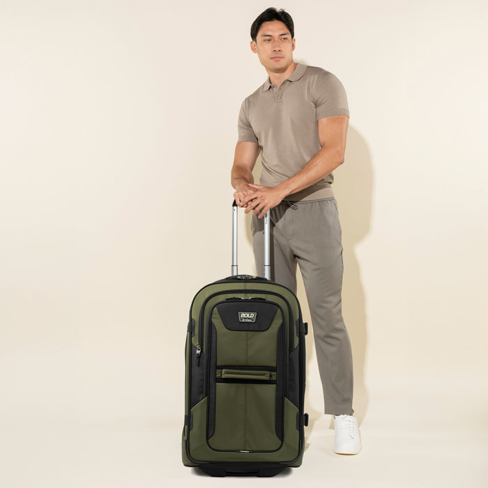 lifestyle view of a male model in a polo shirt and grey pants standing next to the olive black bold by travelpro large check-in expandable rollaboard suitcase, showing its upright vertical scale and extended telescoping handle.