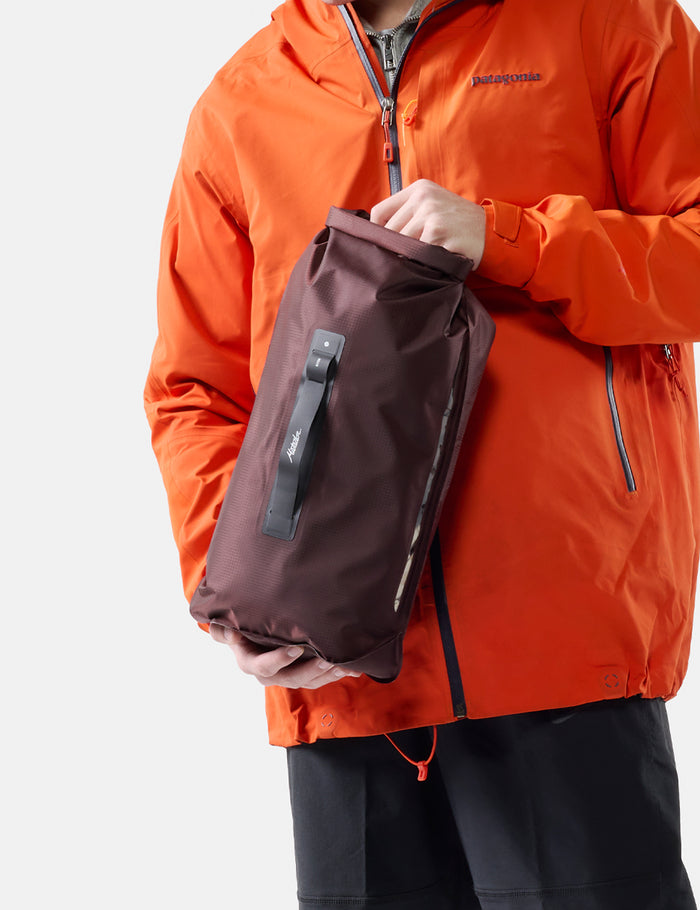 Person wearing an orange jacket holding a garnet dry bag against a white background