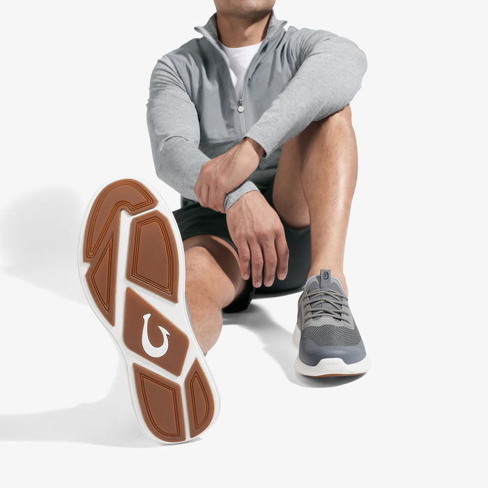 Man wearing gray zip-up jacket, black shorts, and charcoal sneaker with tan sole, shown sitting with shoe sole visible.