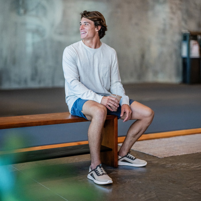 Man wearing light gray long-sleeve shirt and blue shorts with tapa and trench blue colorway, seated on wooden bench.