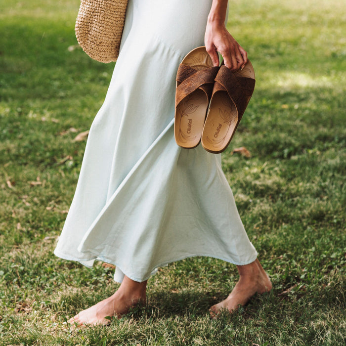 Brown suede slip-on sandals with curved toe design held in hand by person wearing white dress.