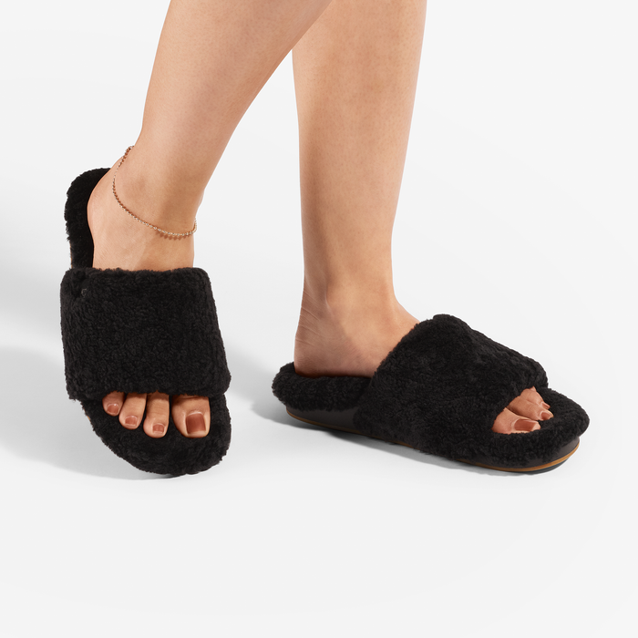 Black fuzzy open-toe slippers with tan soles worn on feet with anklet.