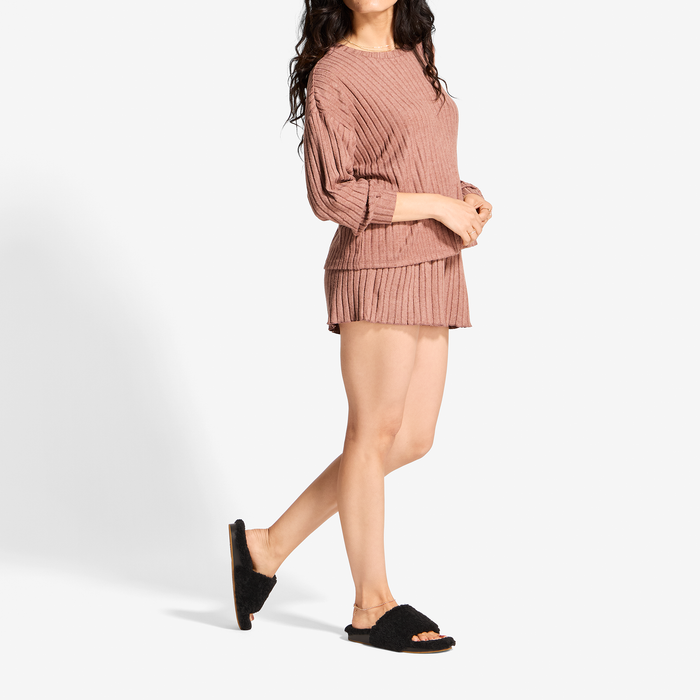 Woman wearing mauve ribbed long-sleeve top and matching pleated mini skirt with black fuzzy slide sandals.