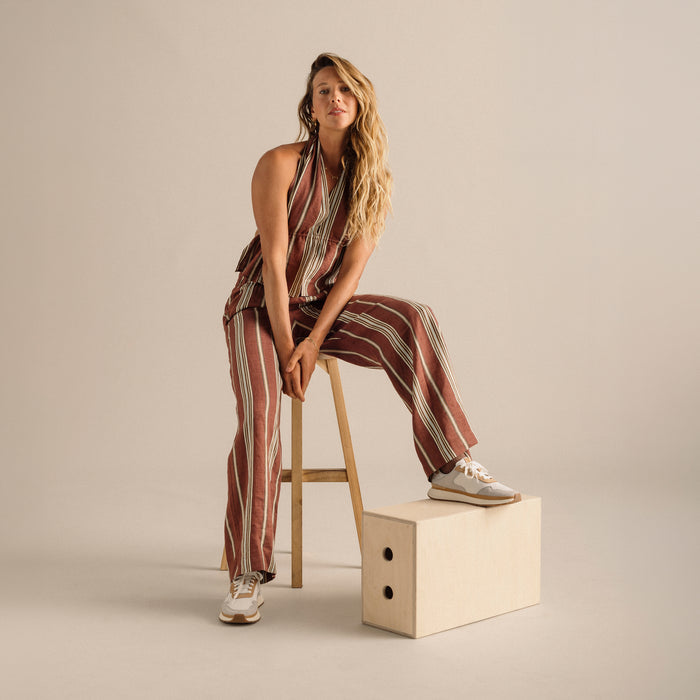 Woman wearing burgundy and cream striped halter jumpsuit with white sneakers, seated on wooden stool.