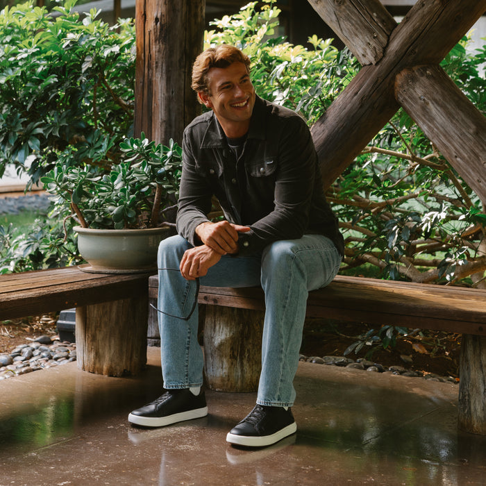 Man wearing black and dark java slip-on sneakers with white sole, seated on wooden bench in garden setting.