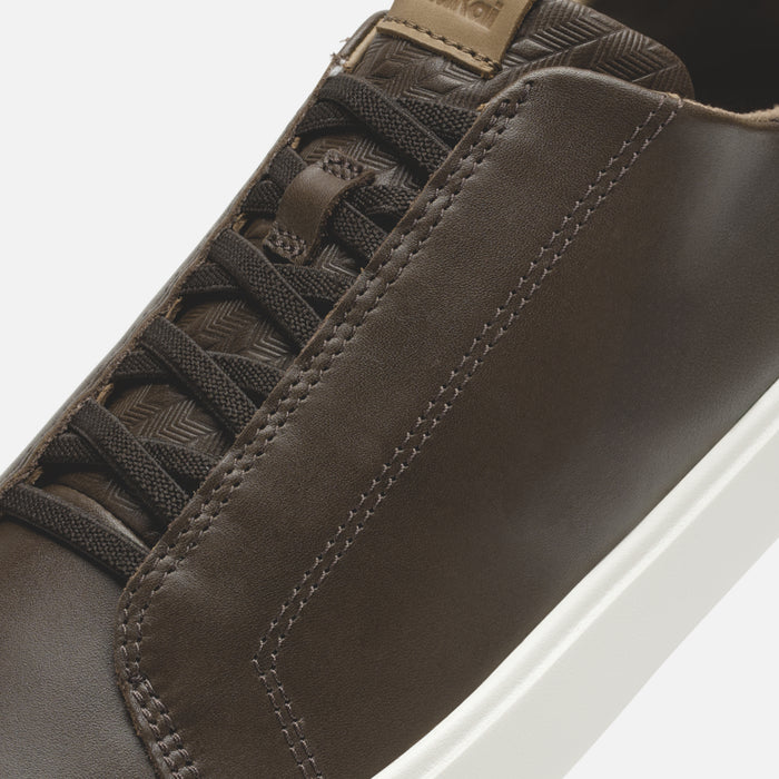 Dark brown leather shoe with tan collar tab, patterned tongue, and white sole.