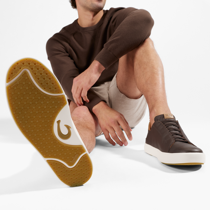 Man wearing dark brown lace-up sneaker with tan sole and geometric tread pattern, paired with brown sweater and tan shorts.