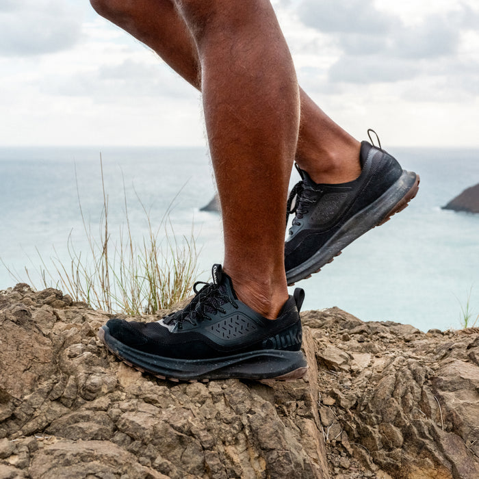 Black lace-up running shoe with textured upper and cushioned sole worn on rocky coastal terrain.