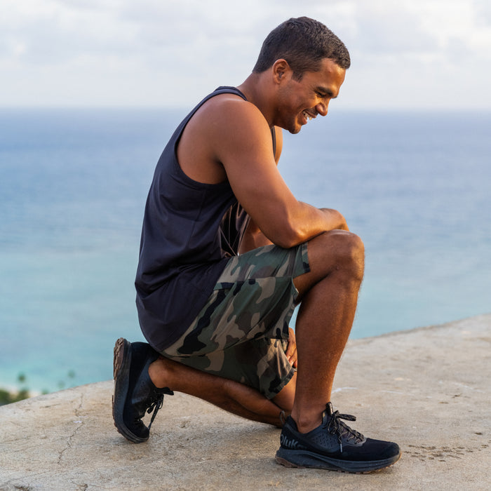 Man wearing black sleeveless tank top and camo-patterned shorts with black sneakers by the ocean.