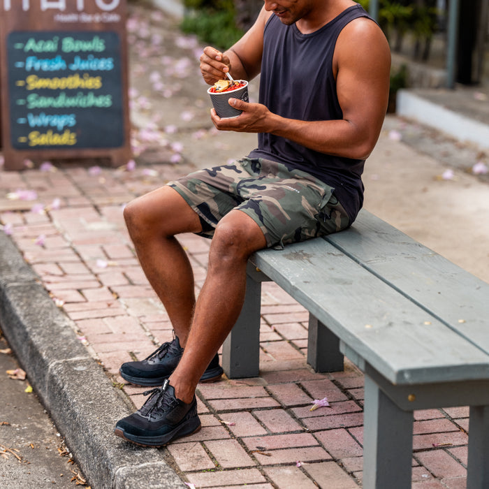 Man wearing black sleeveless tank top and camouflage shorts sitting on a bench.