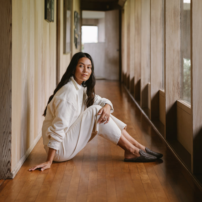 Woman wearing white button-up shirt and dark Java shorts sitting in wooden hallway.