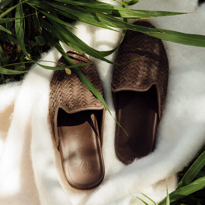Dark brown woven slippers with leather accents on white fabric, surrounded by green plant leaves.