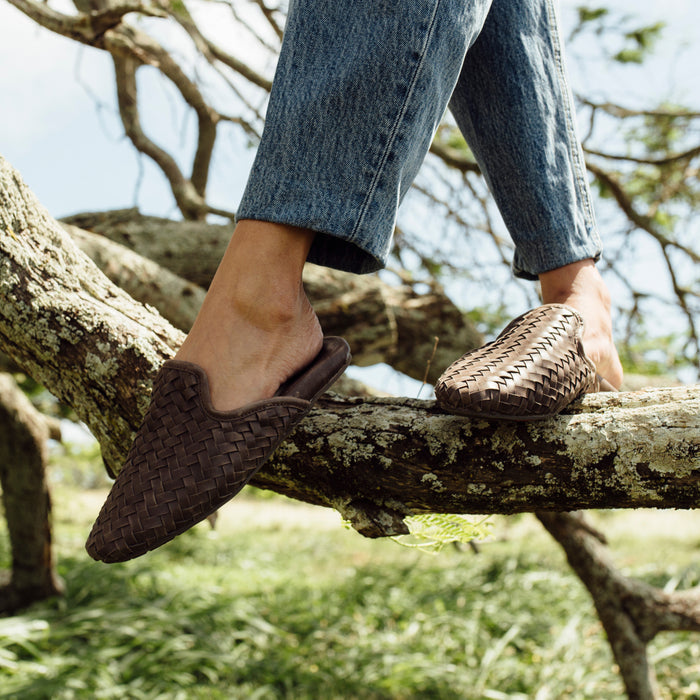 Dark brown woven slip-on shoes worn with cuffed denim jeans on a tree branch outdoors.
