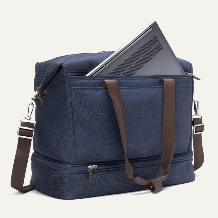 a true navy travelpro platinum elite drop-bottom weekender bag shown with a grey laptop being inserted into the internal padded electronics sleeve, highlighting the bag's organization and tech protection.