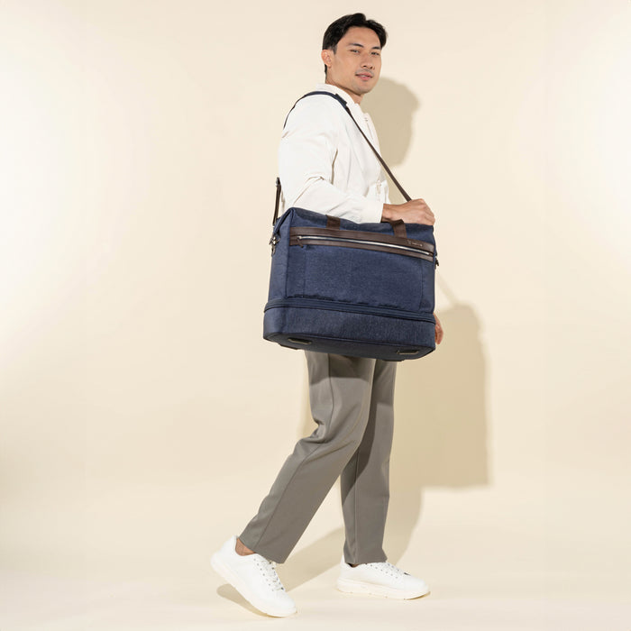 lifestyle shot of a male model walking while carrying the true navy travelpro platinum elite drop-bottom weekender bag using the adjustable shoulder strap, paired with casual white sneakers and tan trousers.