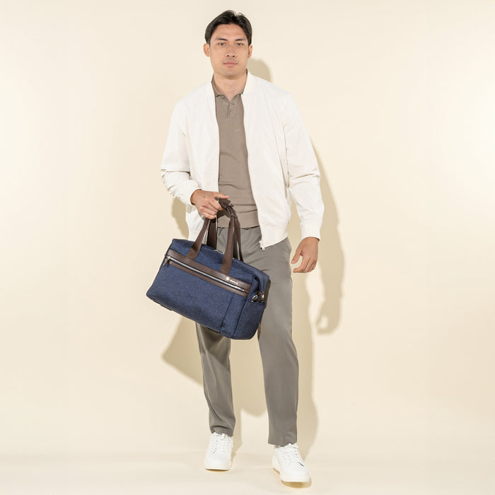 lifestyle shot of a male model in a white jacket and grey trousers, carrying the true navy travelpro platinum elite underseat tote by its leather top handles.