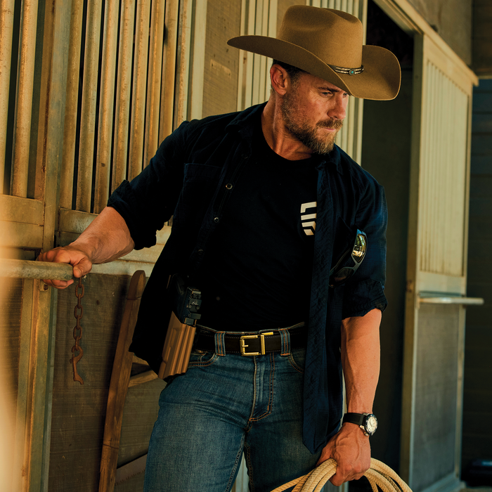 A man in a cowboy hat stands in a barn wearing a black Kore Essentials Ranch Series gun belt with an R1 buckle