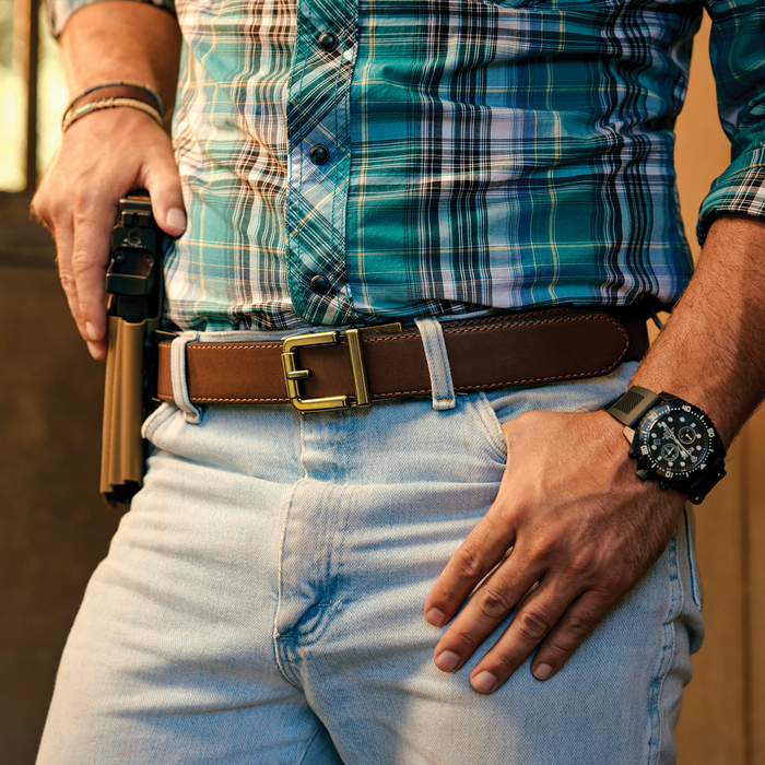 A man stands wearing a brown full grain leather Kore Essentials ranch series gun belt with an R1 brass buckle