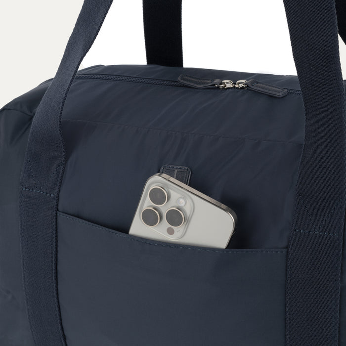 Close-up detail view of the Travelpro Essentials SparePack Foldable Duffel 2.0 in Navy, highlighting the QuickSlip front pocket with a smartphone tucked inside for convenient, quick access to essentials.