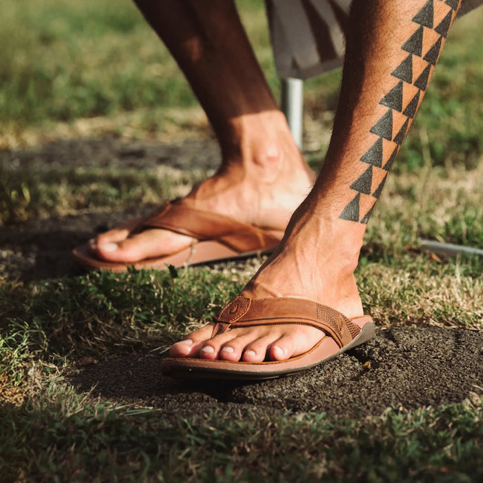 Two men wearing tan leather flip-flops with circular buckle details, standing on grass outdoors.