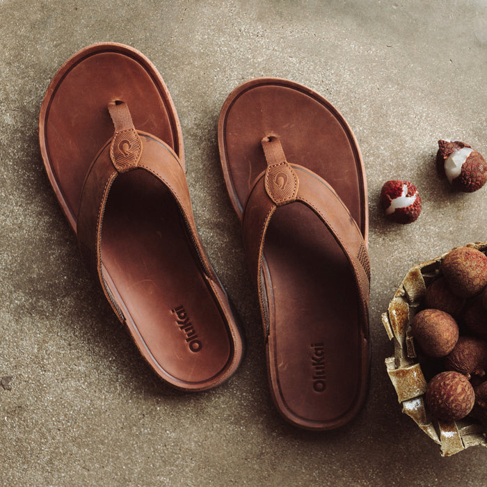 Pair of brown leather thong sandals with circular toe posts and decorative details, displayed on stone surface.