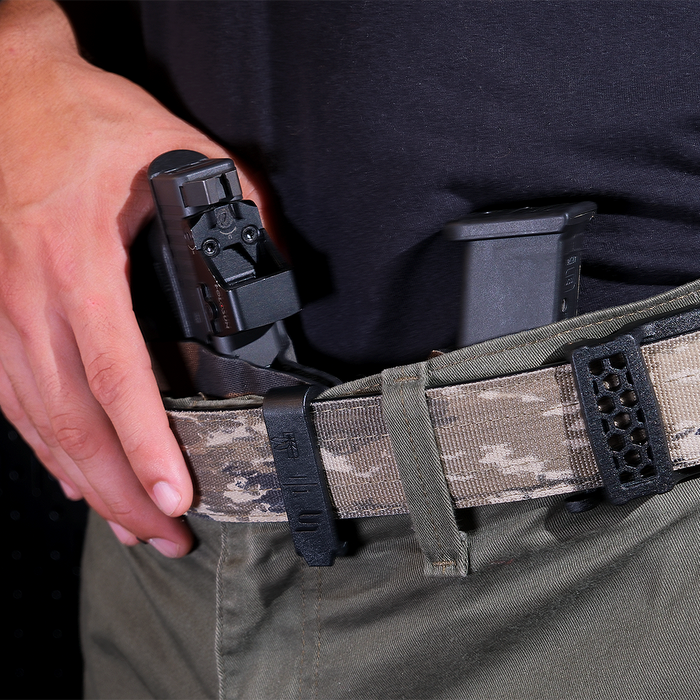 Close-up of a person wearing a camouflage tactical belt with a black quick-release buckle, holding a black handgun in a holster, and carrying a magazine.