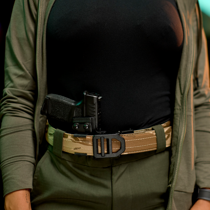 A woman wears a Kore Essentials USA Desert Tiger Stripe EDC gun belt with a firearm holstered at the appendix position.