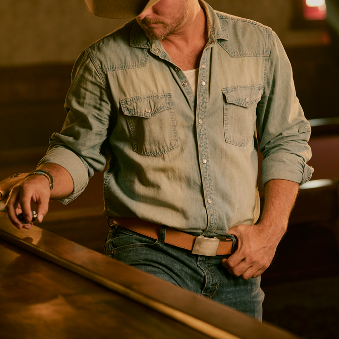 A man wearing denim and a Kore Essentials 1.5" tan buffalo leather EDC gun belt stands at a bar with his head turned down.